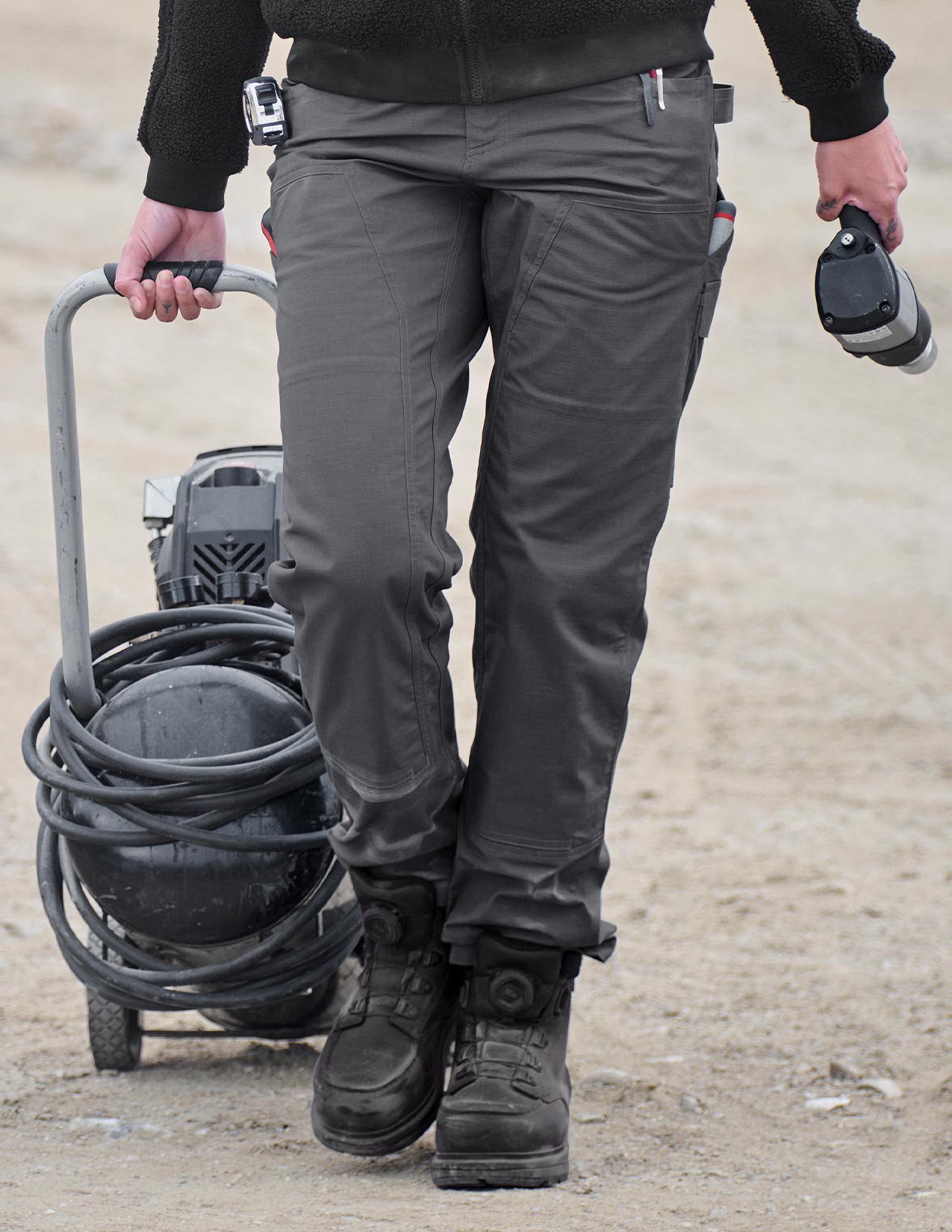 Person wearing dark pants and boots holding a tool and hose on a dirt ground.