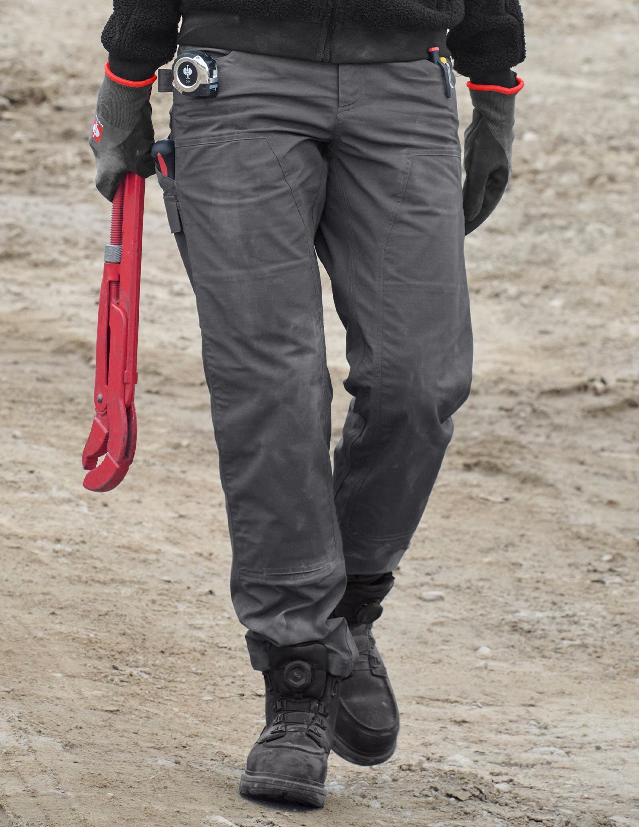 Person wearing gray pants and black boots, holding red tools on a dirt ground.