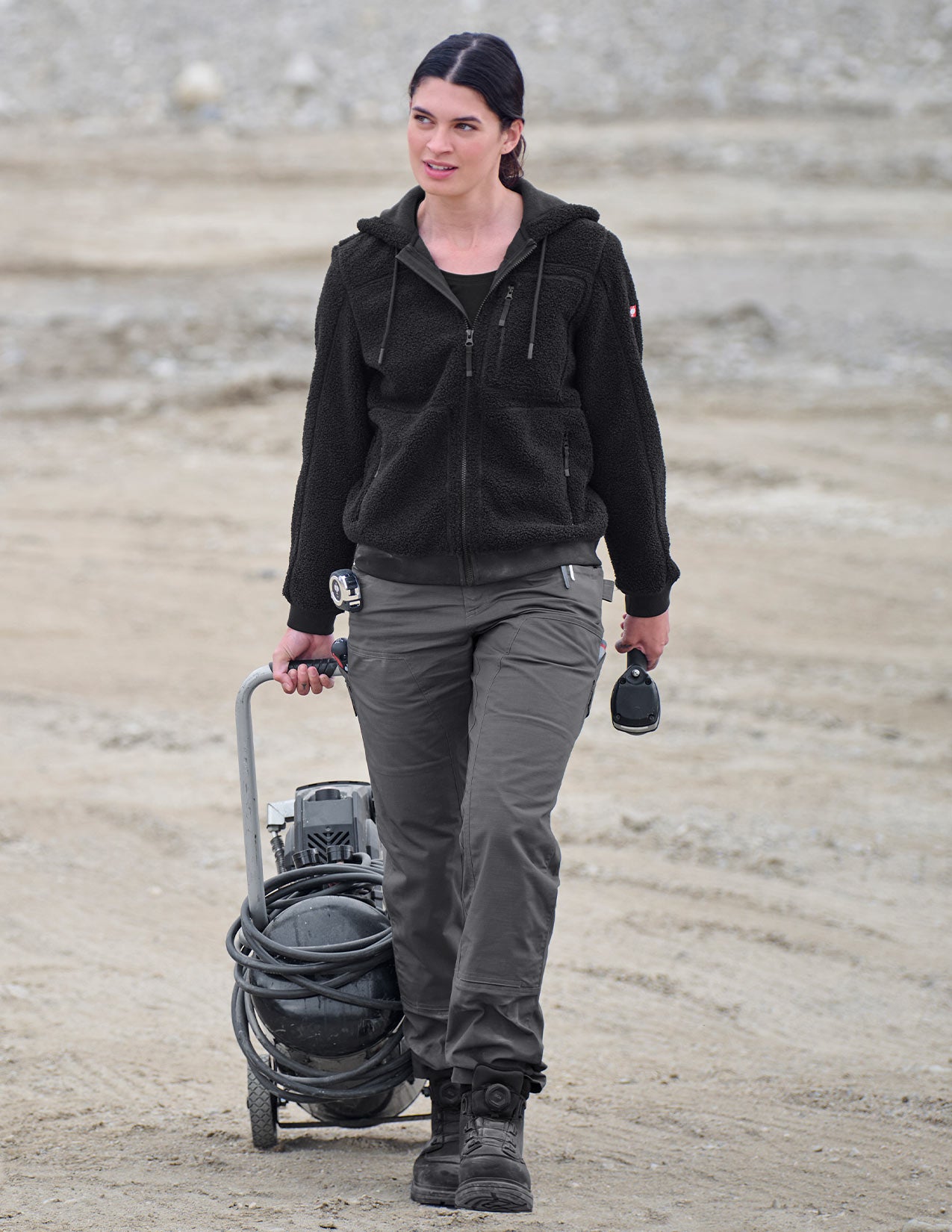 Person in black jacket and gray pants walking with equipment on a dirt path