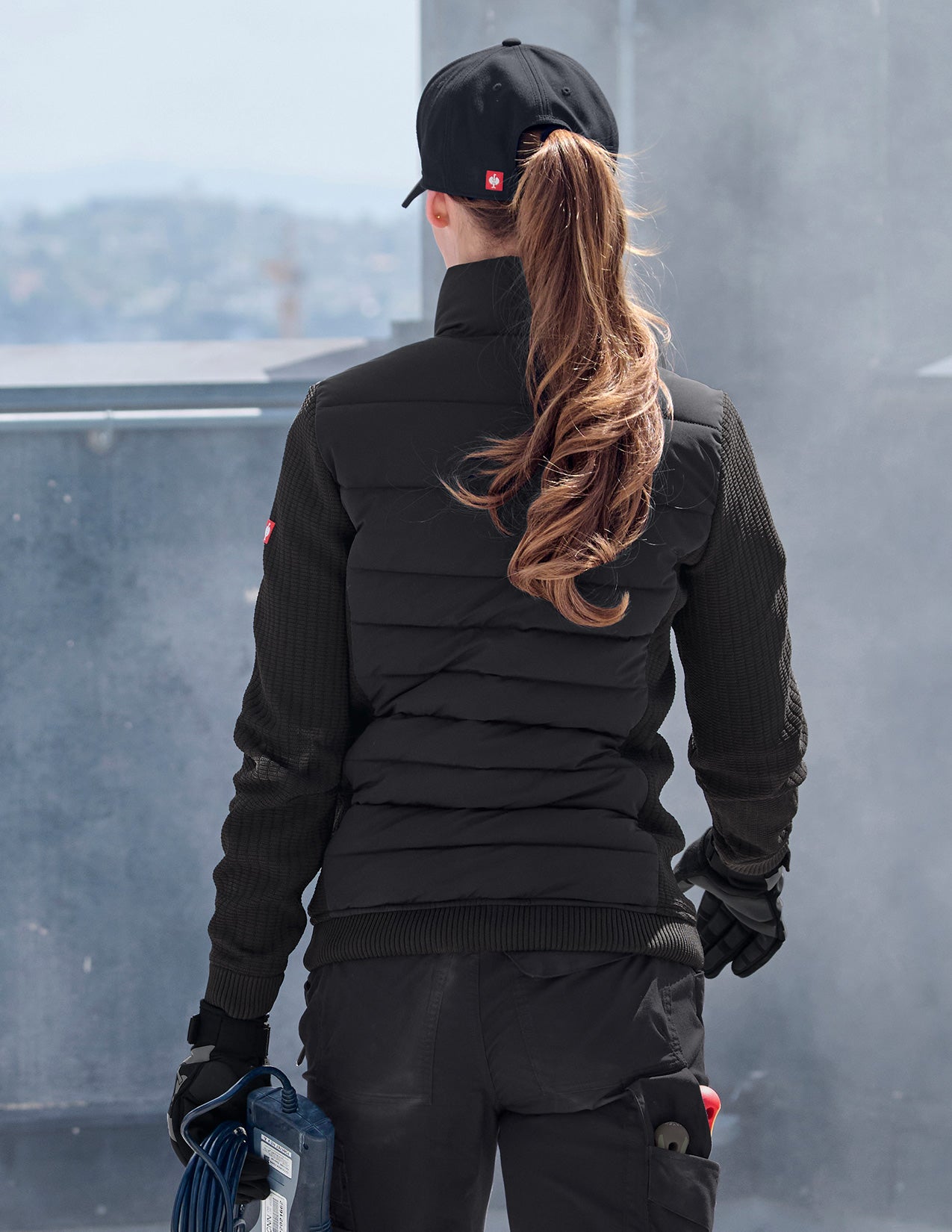 Person wearing a black puffer jacket and cap, standing on a rooftop with a cityscape view.