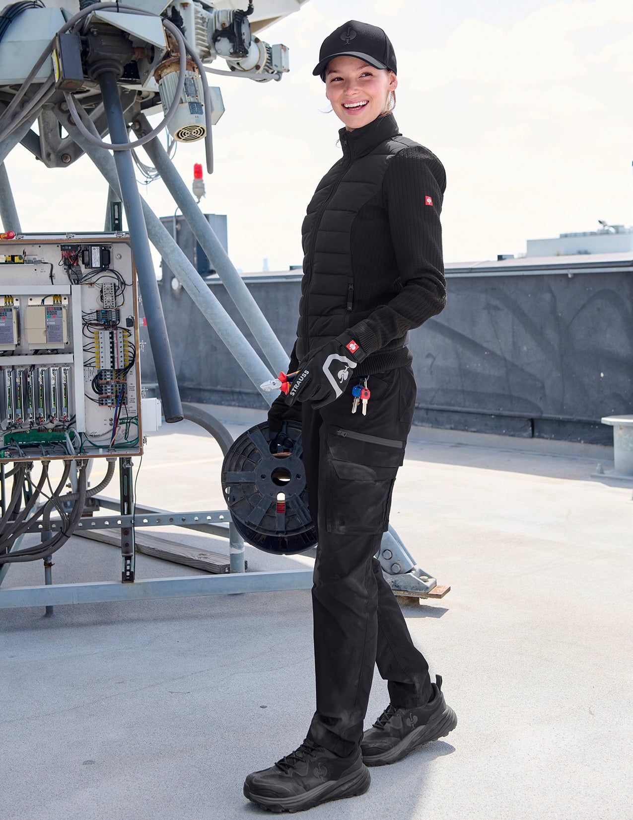 Person wearing a black puffer jacket on a rooftop with technical equipment in the background.