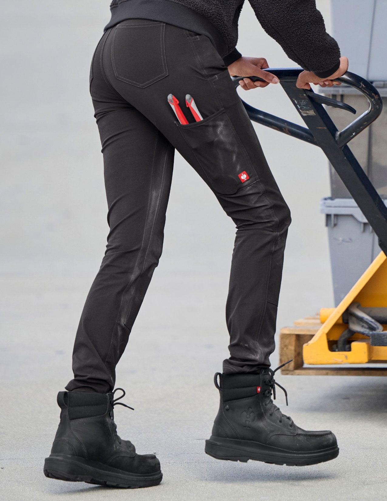 Person wearing black pants with red accents and black boots, using a pallet jack.