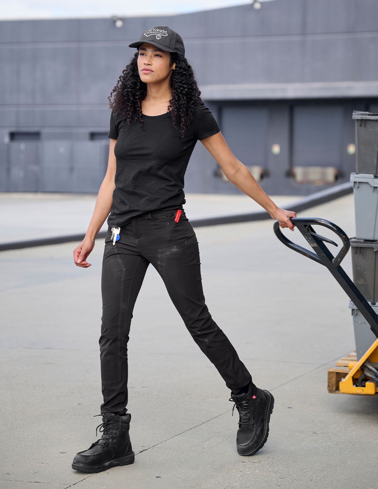 Person wearing a black outfit with a cap, standing outdoors near a building.
