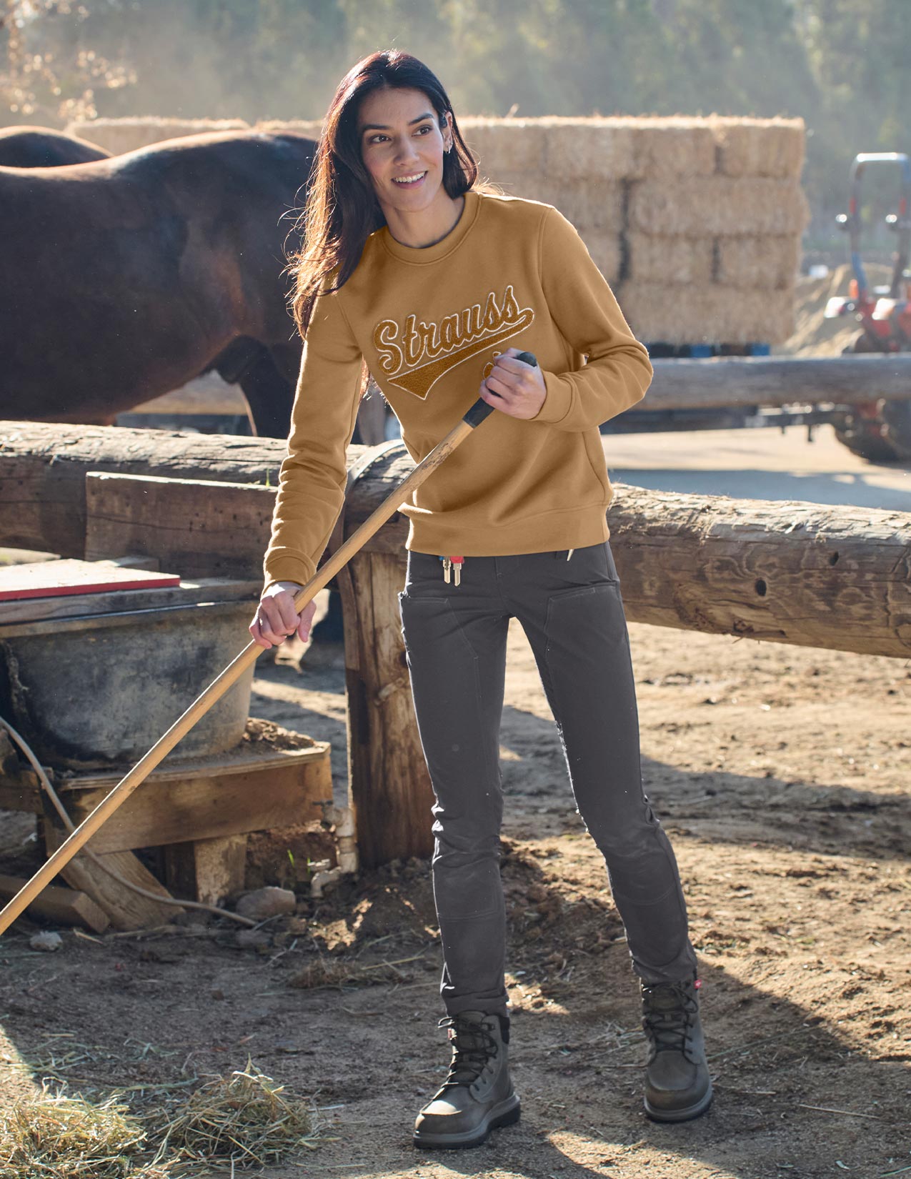 Woman in a brown sweater with 'Strauss' logo standing next to a horse in a barn setting.