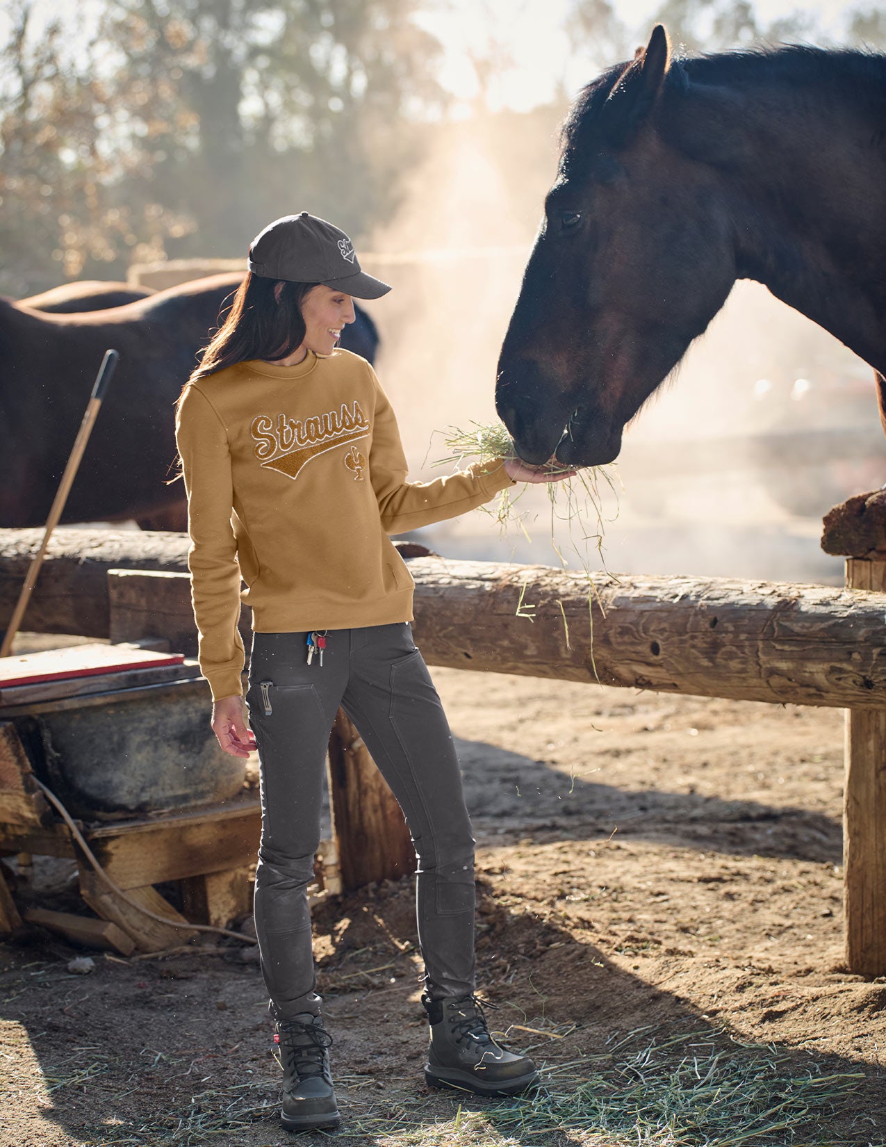 Woman in a brown sweatshirt and gray pants feeding a horse in a barn setting.