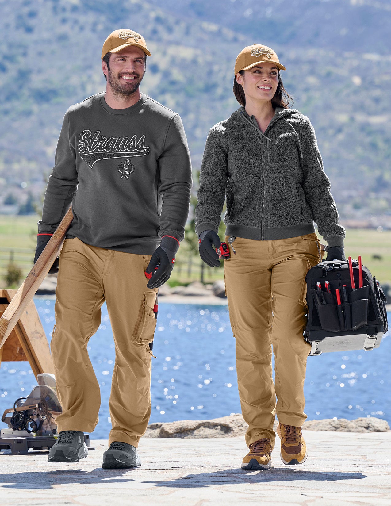 Two people in outdoor work attire standing on a dock with mountains in the background.