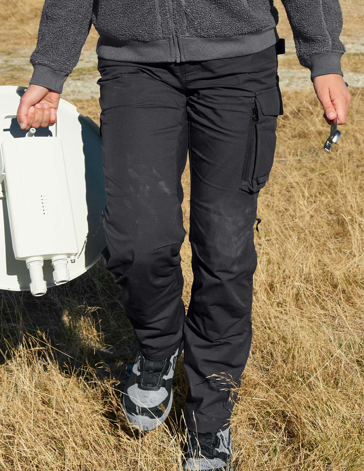 Person wearing black cargo pants walking through a field with a white device.