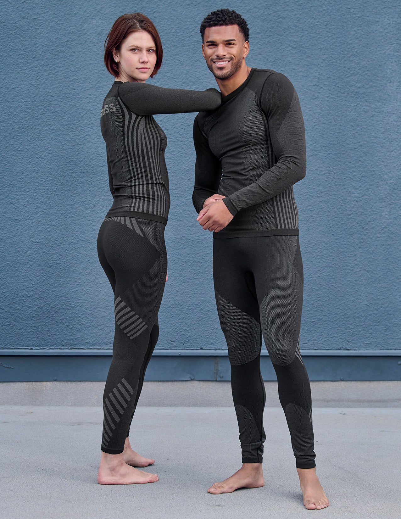 Two people wearing black thermal pants and longsleeves standing against a blue wall.