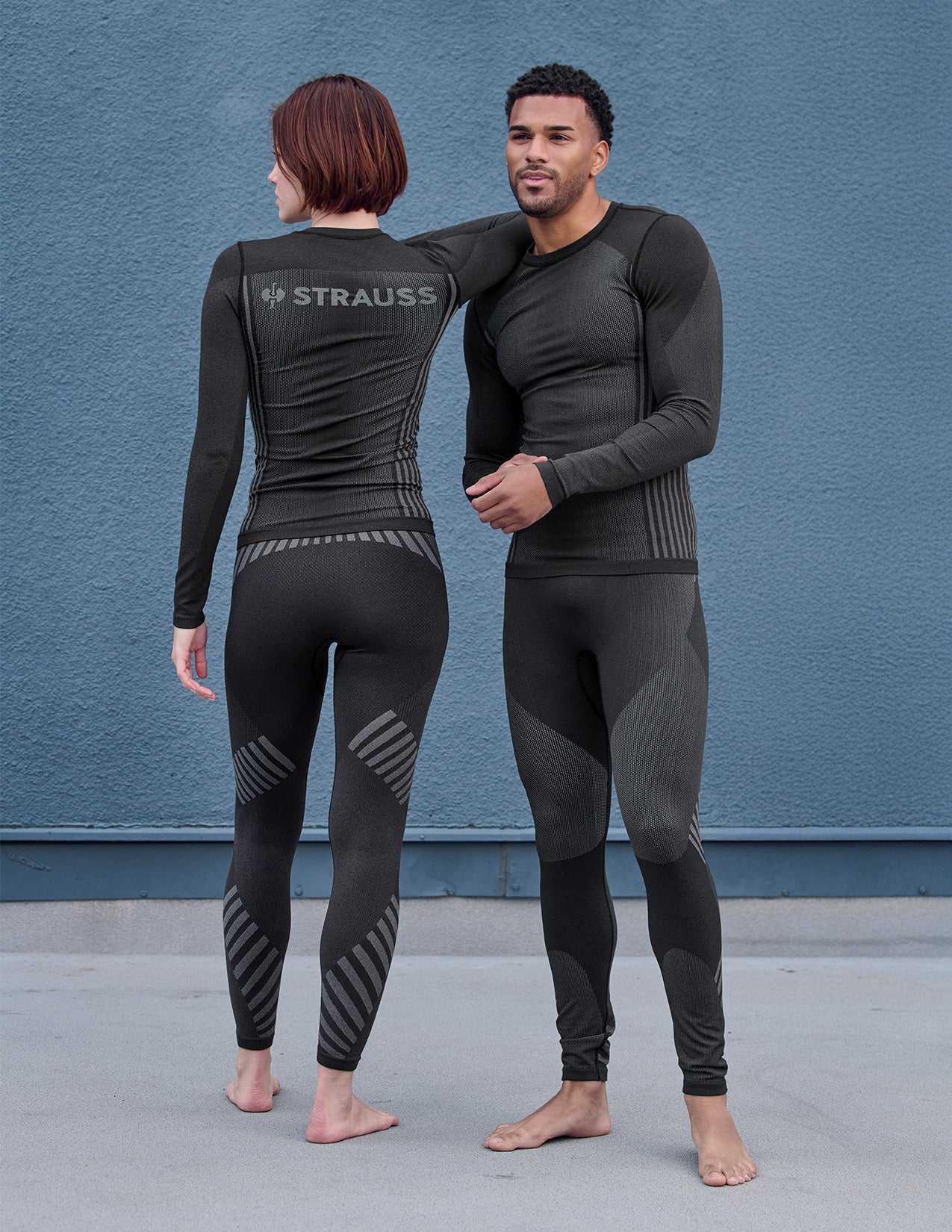 Two people wearing black Strauss thermal pants and longsleeves against a blue wall.