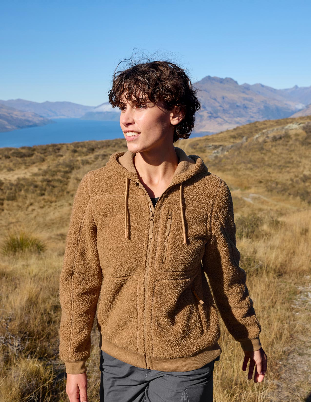Women wearing a brown jacket standing in a scenic outdoor setting with mountains and water.