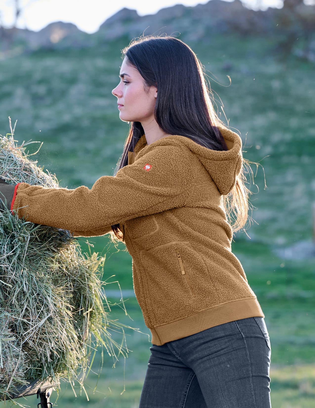 Woman in a brown jacket holding a bundle of hay in a field