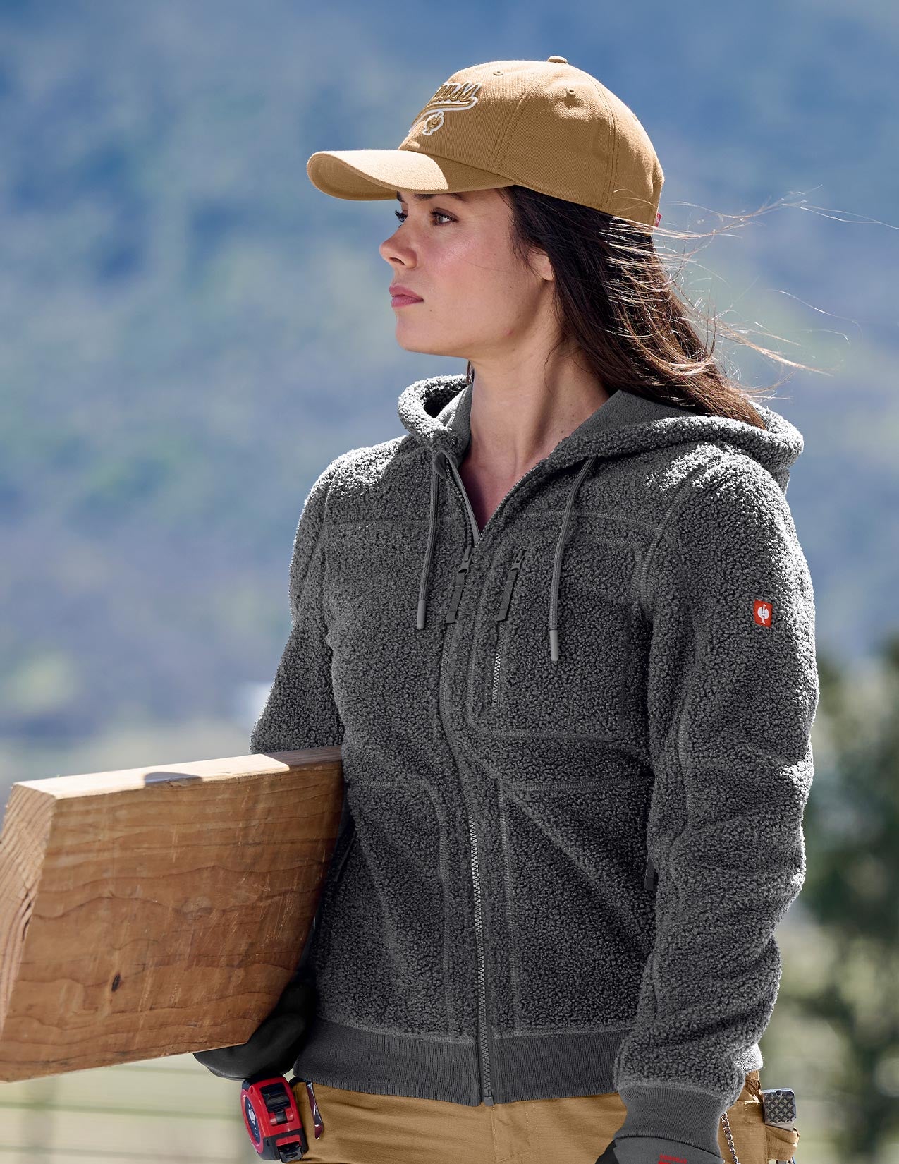 Woman wearing a gray jacket and brown cap, holding a log with a mountainous background