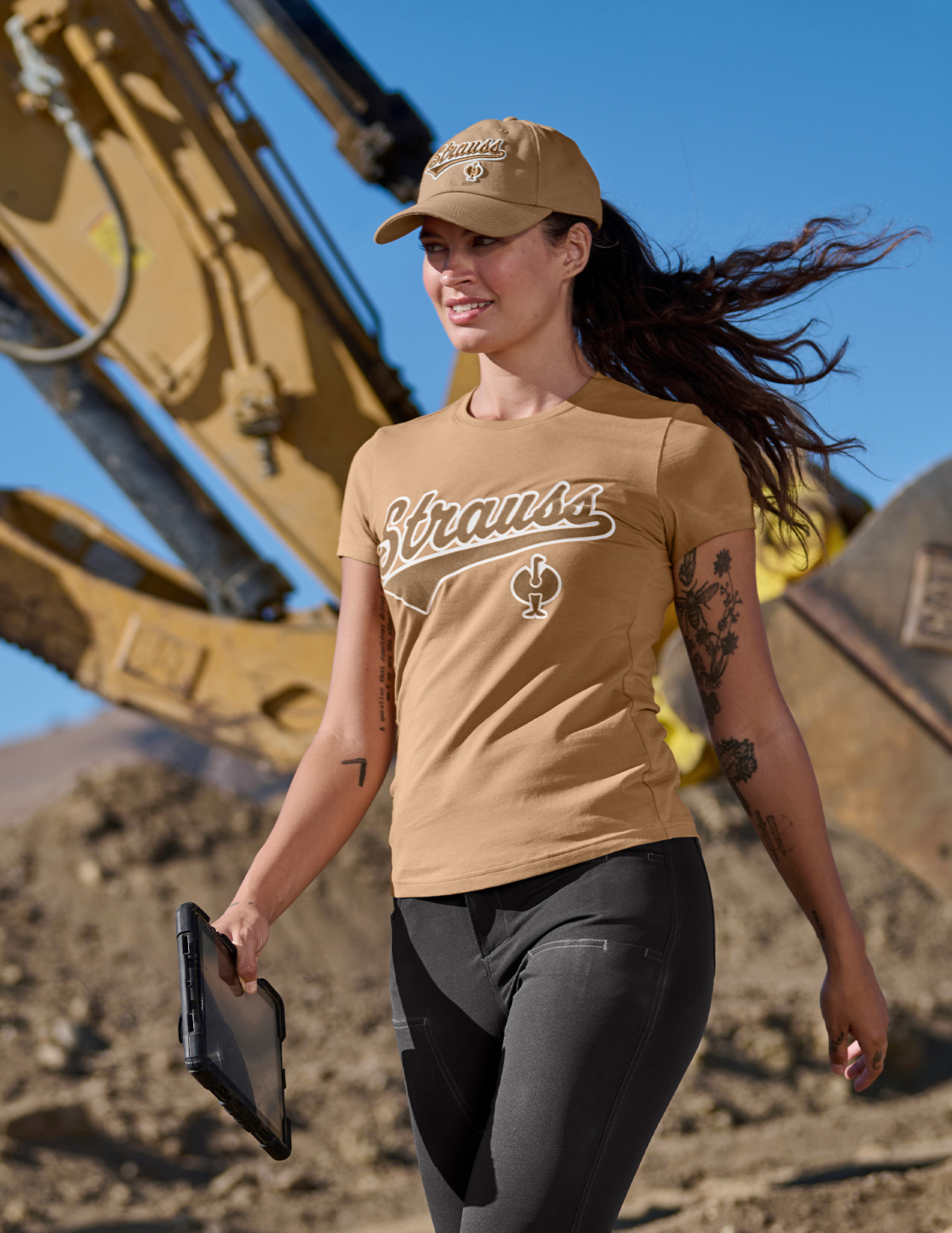 Woman wearing a shirt and cap with a logo, standing in front of construction equipment.