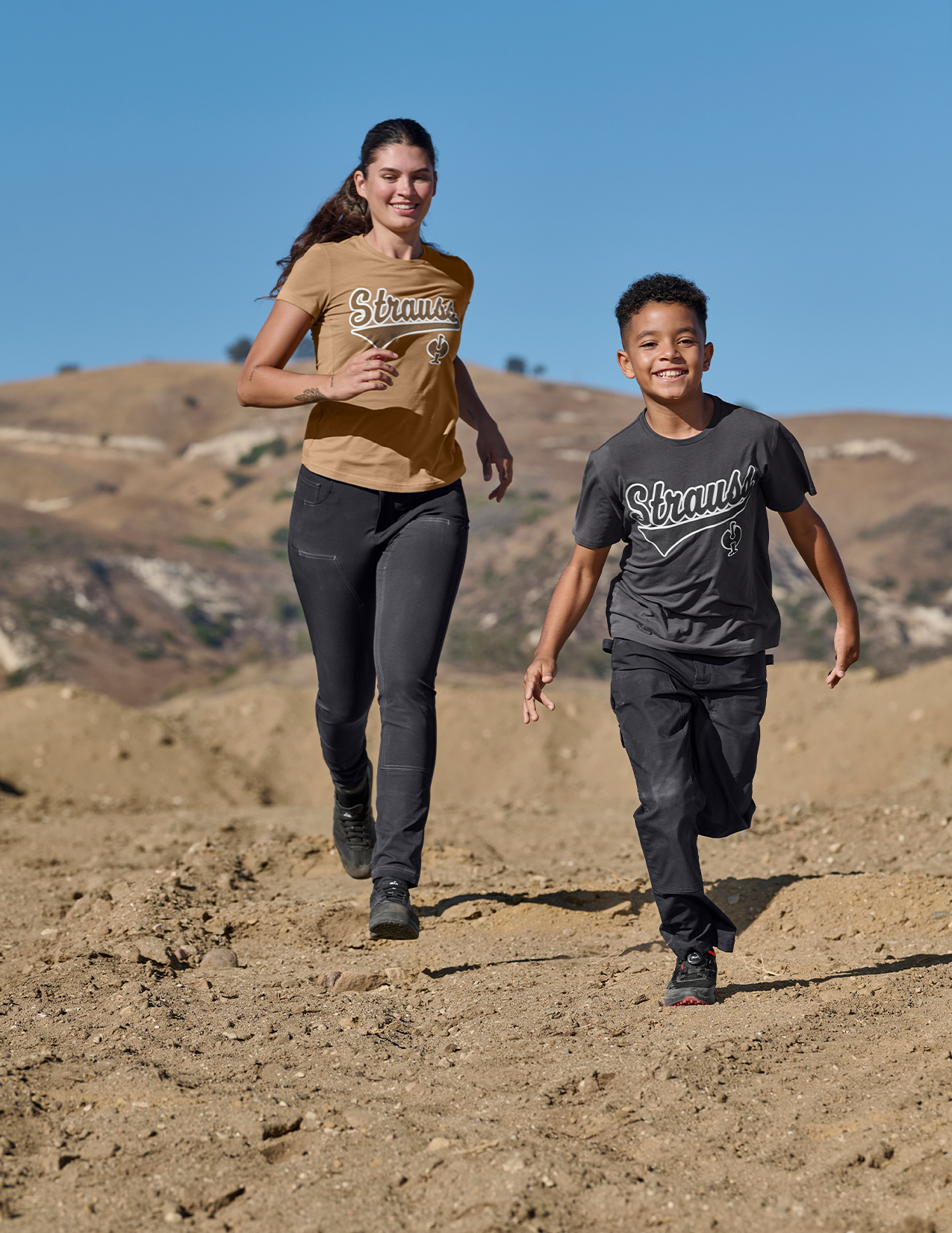 Two people running on a dirt path with a clear blue sky and hills in the background.