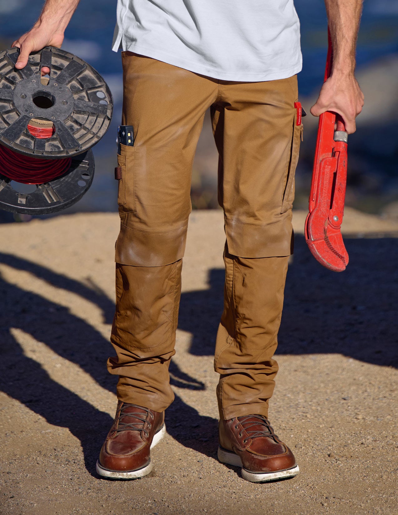 Person wearing brown pants and holding a red tool and spool of wire.
