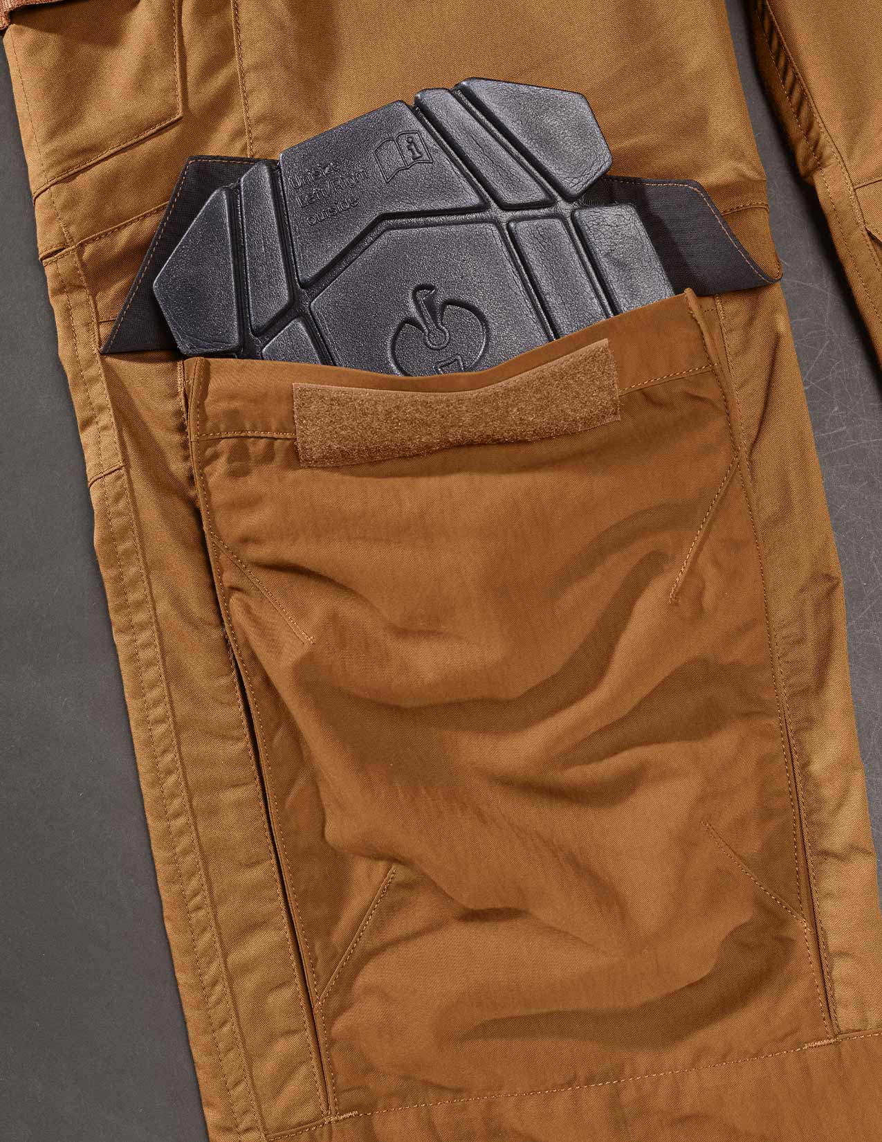 Brown pants with a black kneepad in the pocket on a gray background
