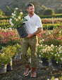 Man holding a potted plant in a garden setting with flowers and mountains in the background