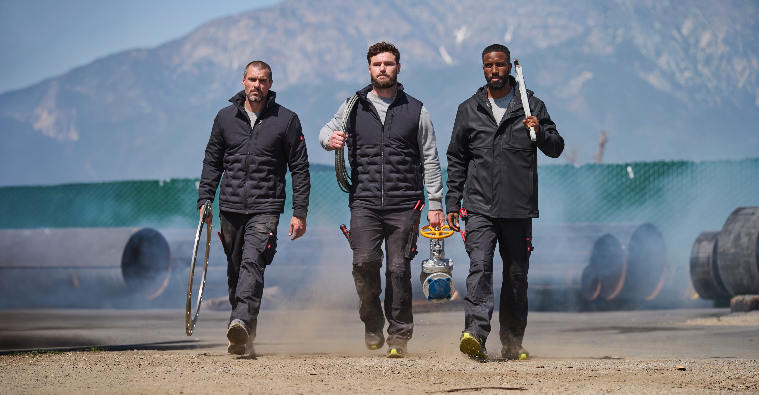 Three men walking outdoors on a sandy surface with mountains and water in the background. One man wears a black puffer vest over a gray long-sleeve shirt and dark pants, carrying a coiled hose.