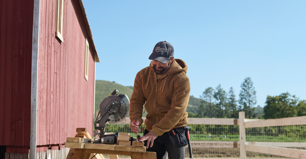 A man wearing a brown sherpa fleece zip-up hoodie, a dark gray and white STRAUSS baseball cap, and black pants is working outdoors next to a red barn, using a miter saw.