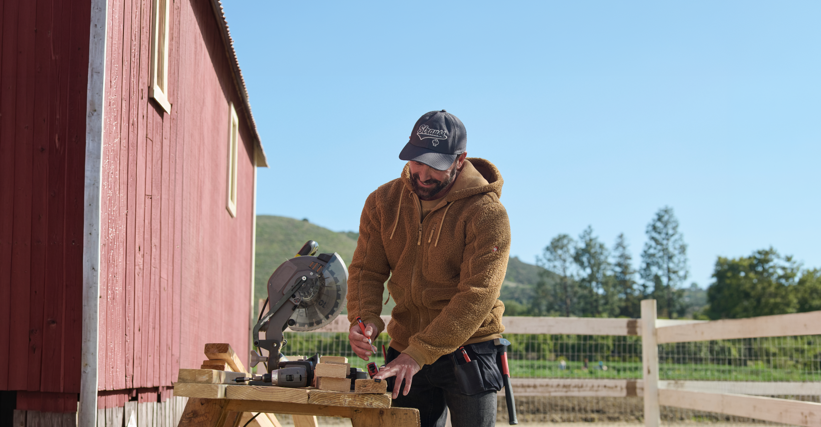 A man wearing a brown sherpa fleece zip-up hoodie, a dark gray and white 'Kings of NY' baseball cap, and black pants is working outdoors next to a red barn, using a miter saw.