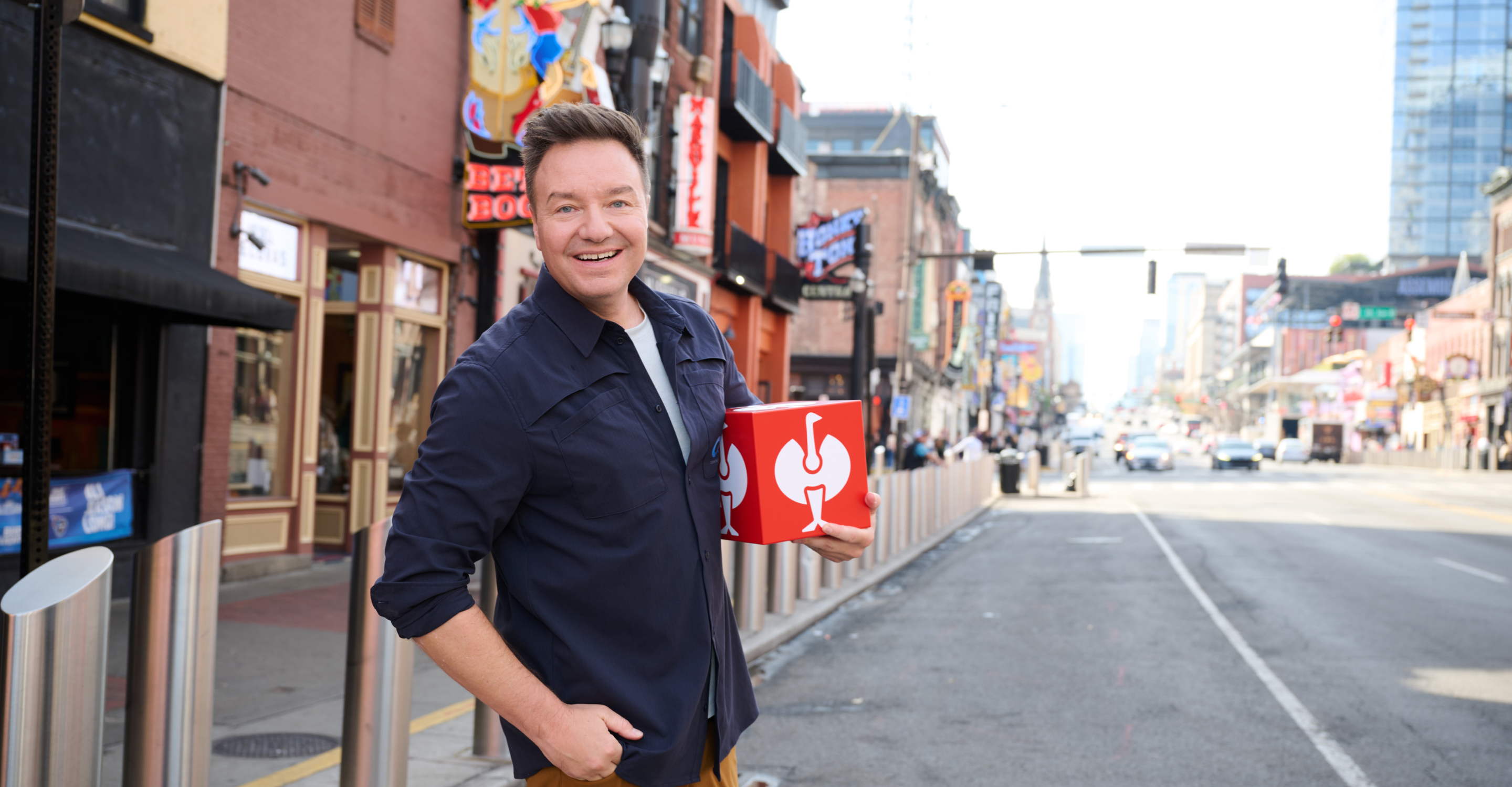Henning Strauss standing on a street in Nashville while holding a red box with a white STRAUSS logo. The background shows a blurred urban street scene with buildings, storefronts, and road markings.