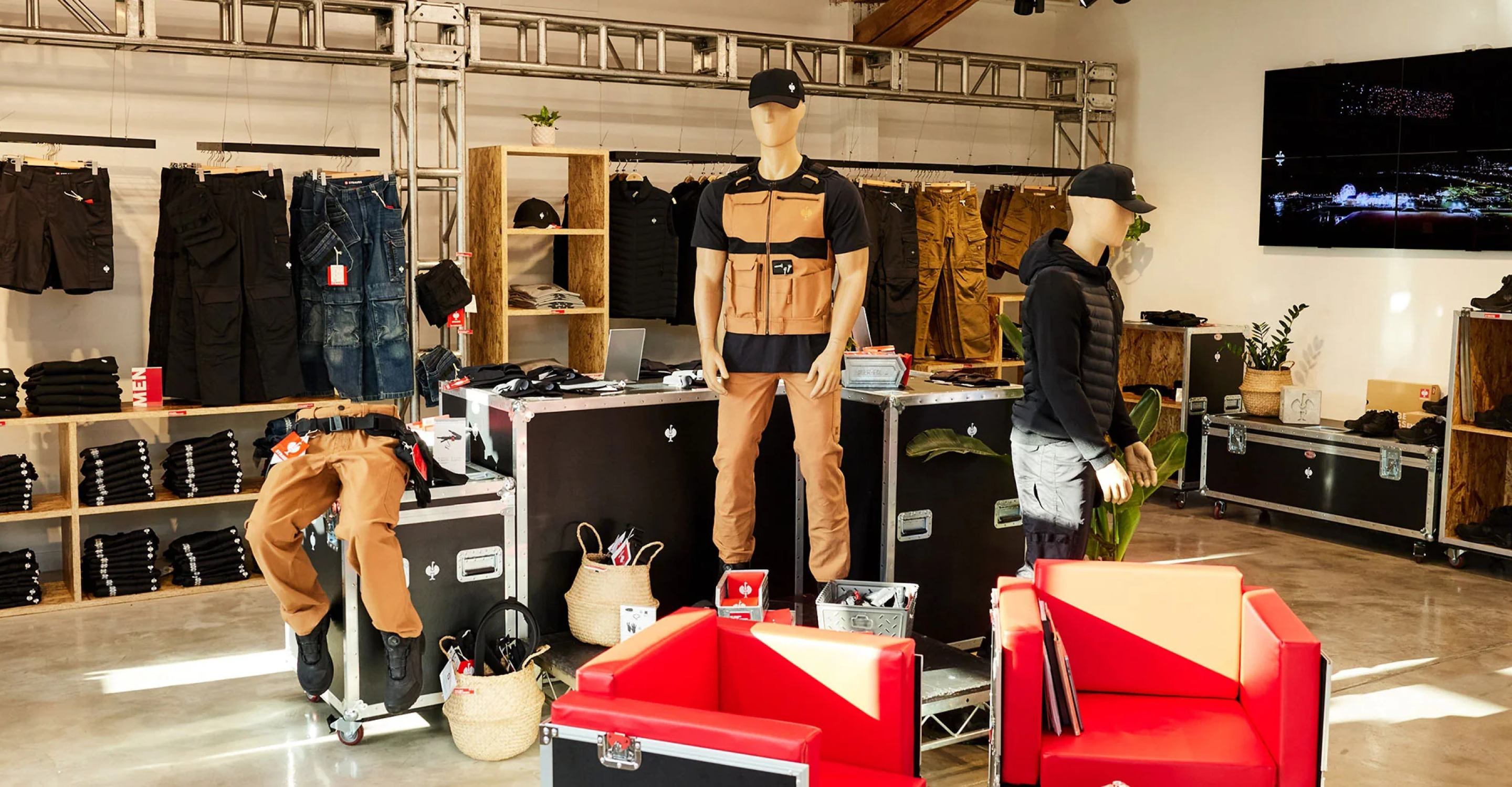 A retail display room showcasing outdoor and workwear apparel, featuring mannequins and a real person modeling tan and black utility gear, with red chairs in the foreground and racks of clothing in the background.