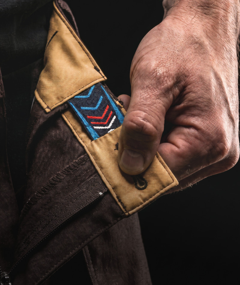 A close-up of a person's hand pulling open a dark brown jacket to reveal a tan interior pocket with a colorful embroidered chevron patch in blue, red, white, and black.