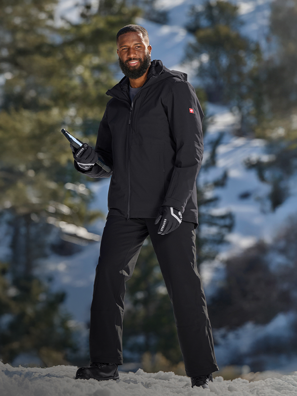 Person in black winter clothing holding a bottle in a snowy landscape with trees and mountains.