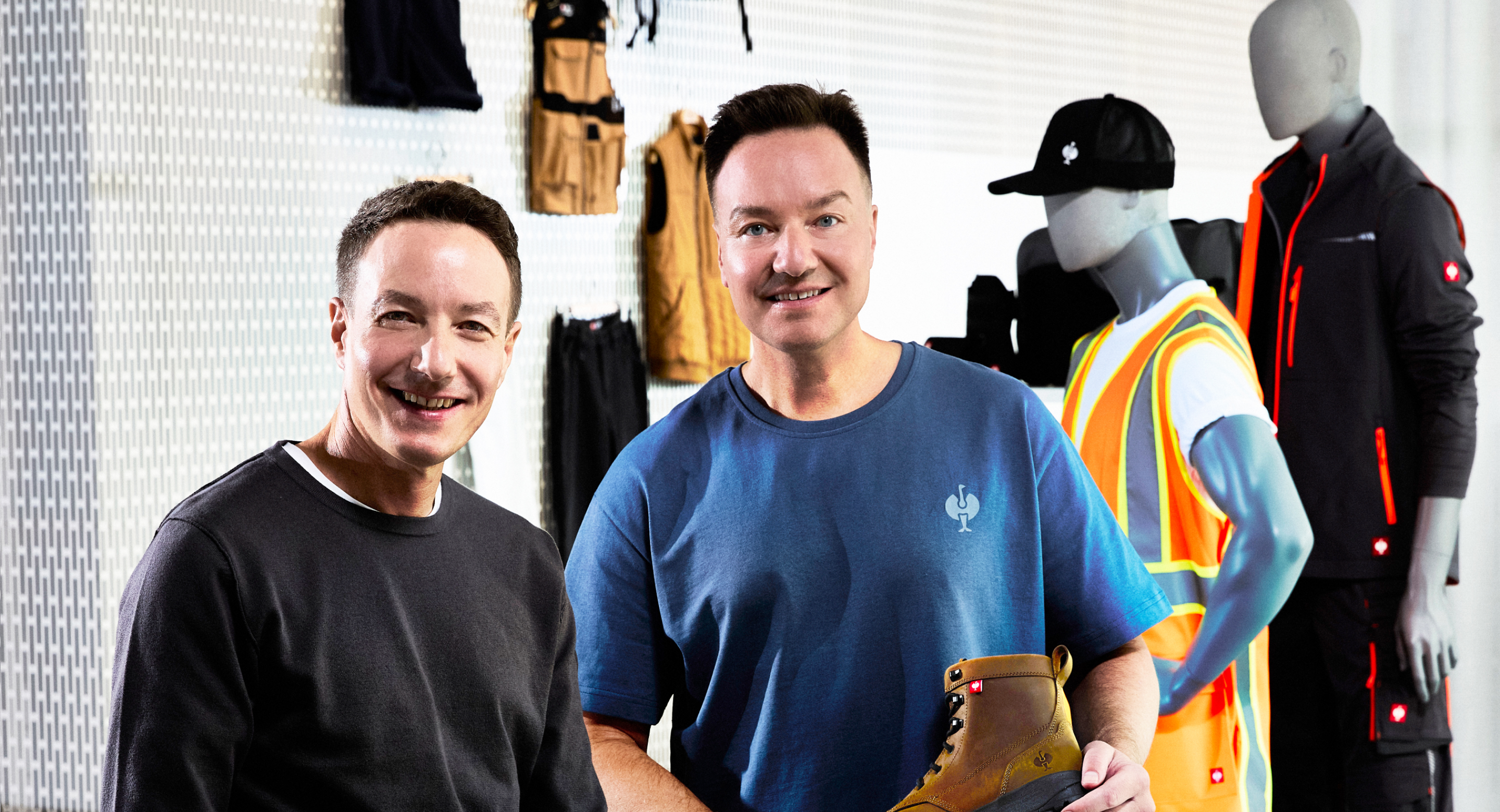 Two men standing in a retail or workshop setting, with one holding a tan work boot. Mannequins and other apparel are visible in the background against a white wall.