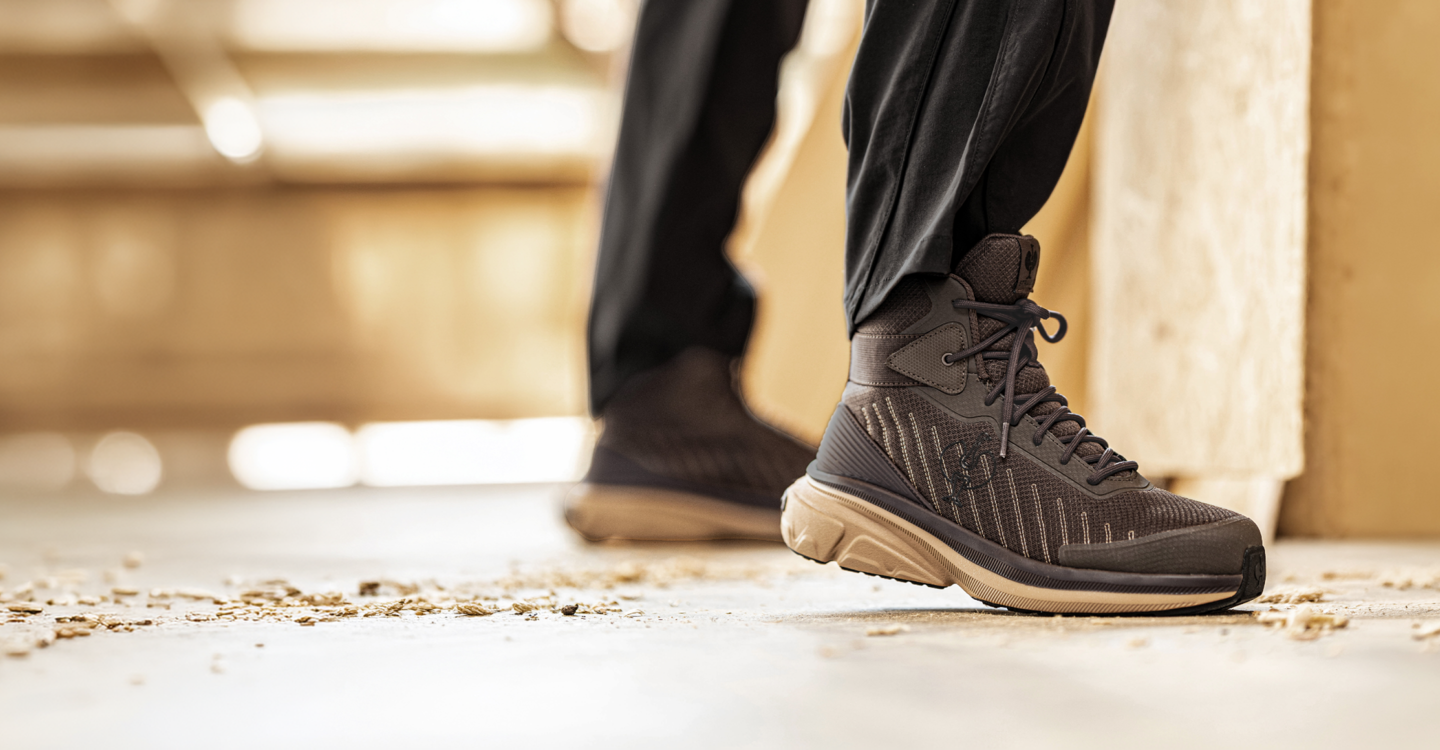 A close-up of a person wearing a dark brown and black work boot with black pants, standing on a concrete floor with a blurred background.