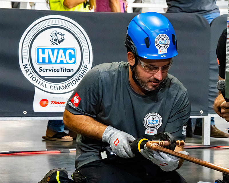 A man wearing a blue hard hat, safety glasses, and gray gloves kneels down in an indoor event space, working on a copper pipe fitting. A large banner in the background reads 'HVAC ServiceTitan National Championship'.