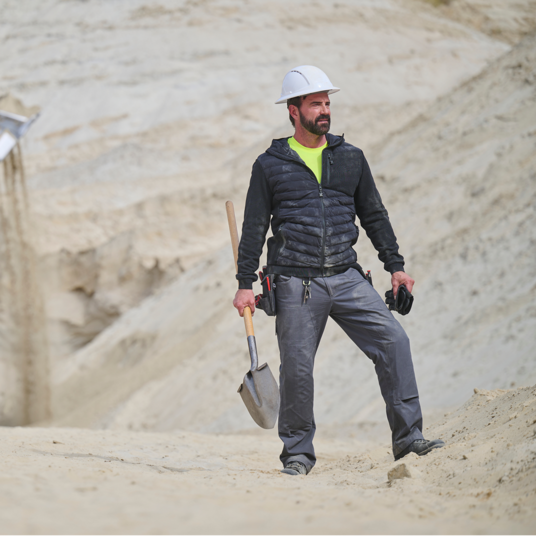 Man in construction attire with a hard hat and tools standing on a construction site.