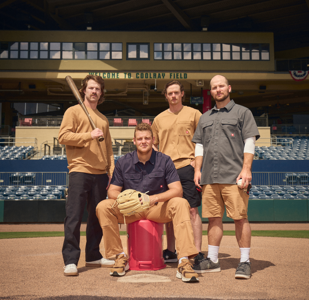 Four men posing together on a baseball field, wearing a coordinated set of tan and brown baseball apparel from STRAUSS. One man holds a wooden baseball bat, another holds a baseball, and one wears a glove.