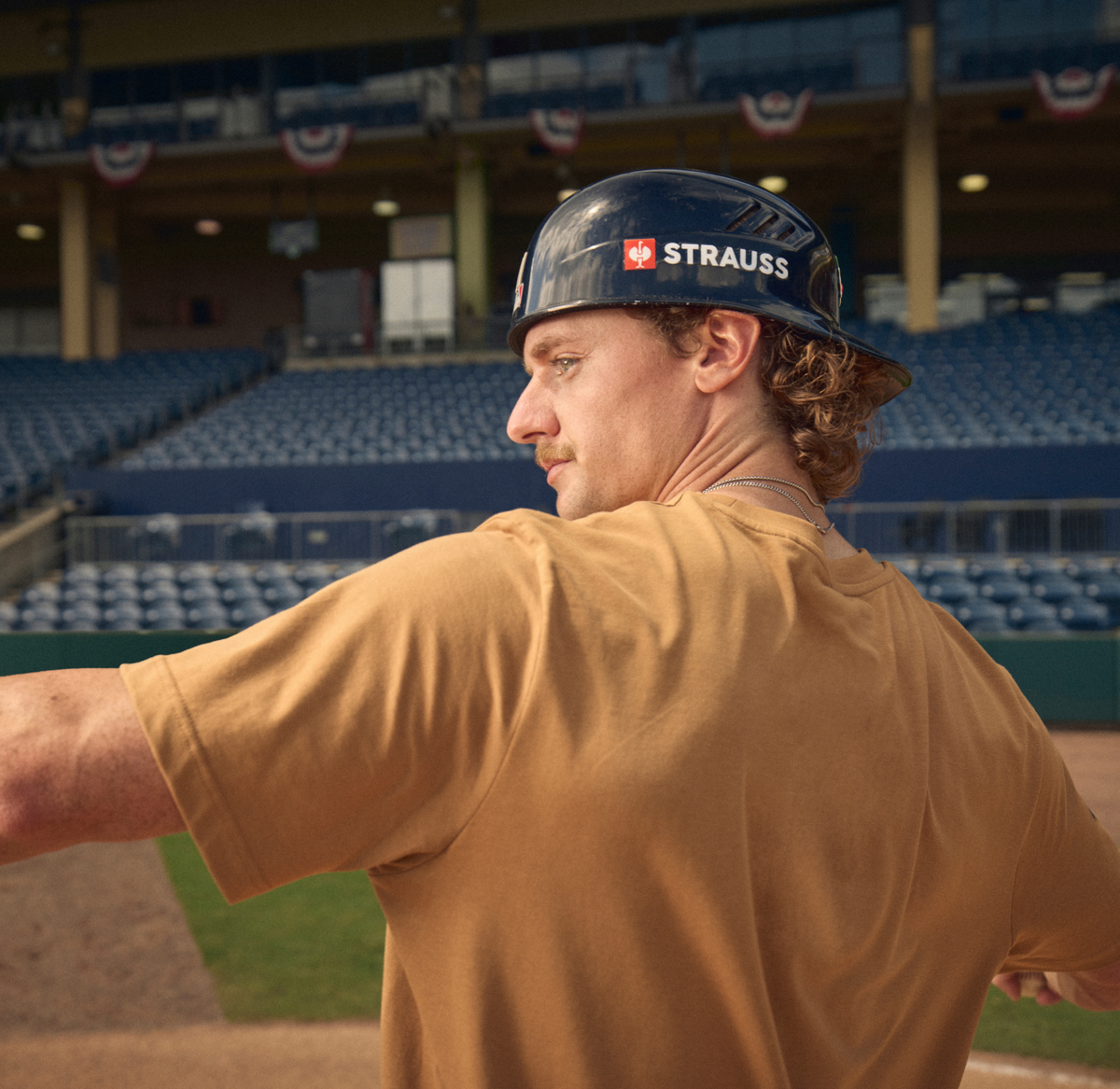 A man in a tan jersey and a black Strauss batting helmet, shown from behind in a stadium with empty blue seats in the background.
