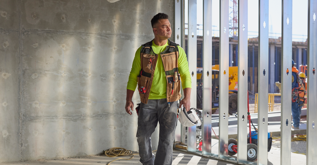 Nick Swisher wearing a bright yellow long-sleeve shirt, an almondbrown tool vest, and gray work pants walks through an open steel frame structure on a construction site.