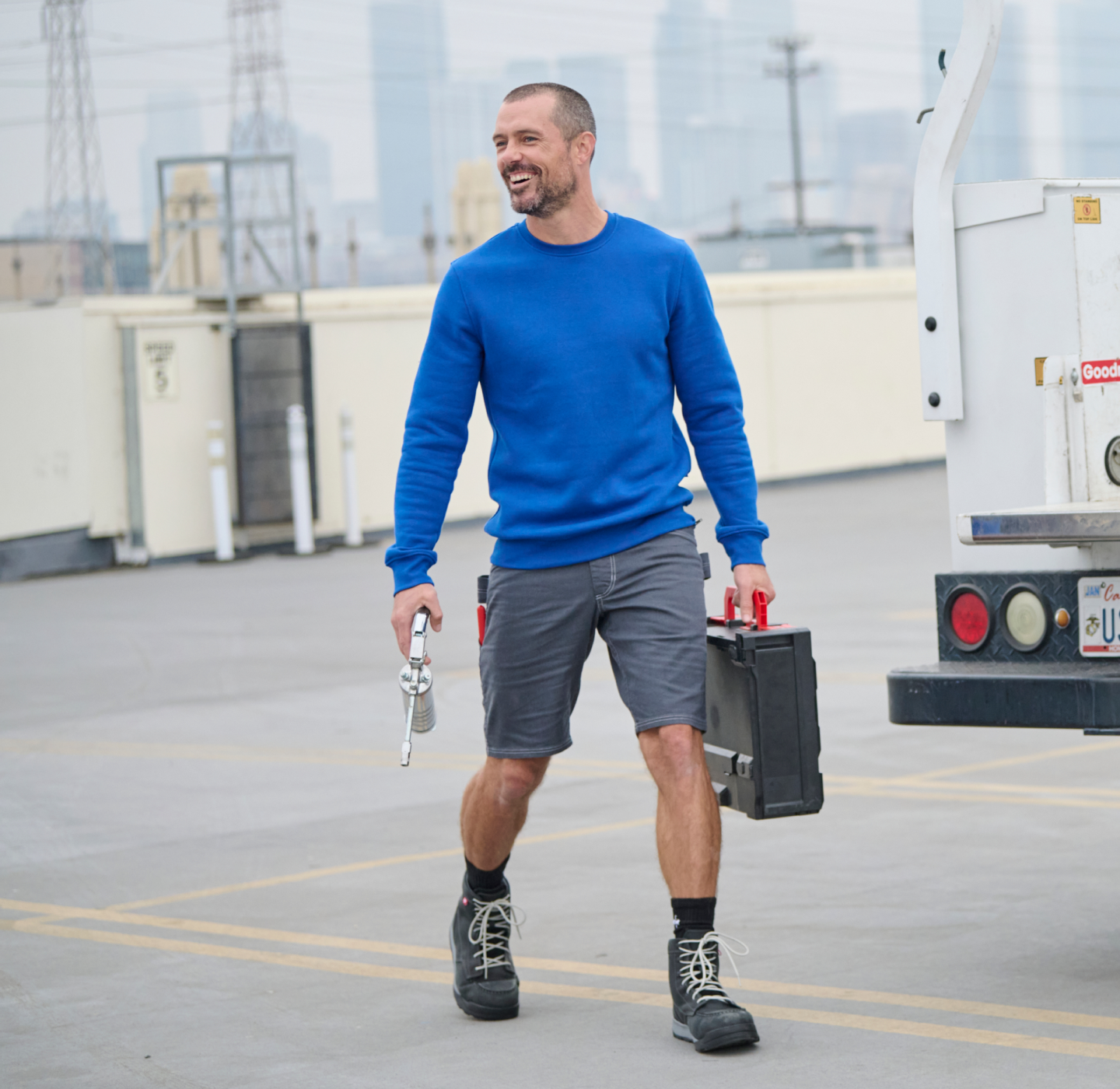 A man in a blue sweater, gray shorts, and black boots smiles while walking in a parking lot, holding a black toolbox and a wrench near a white utility truck.