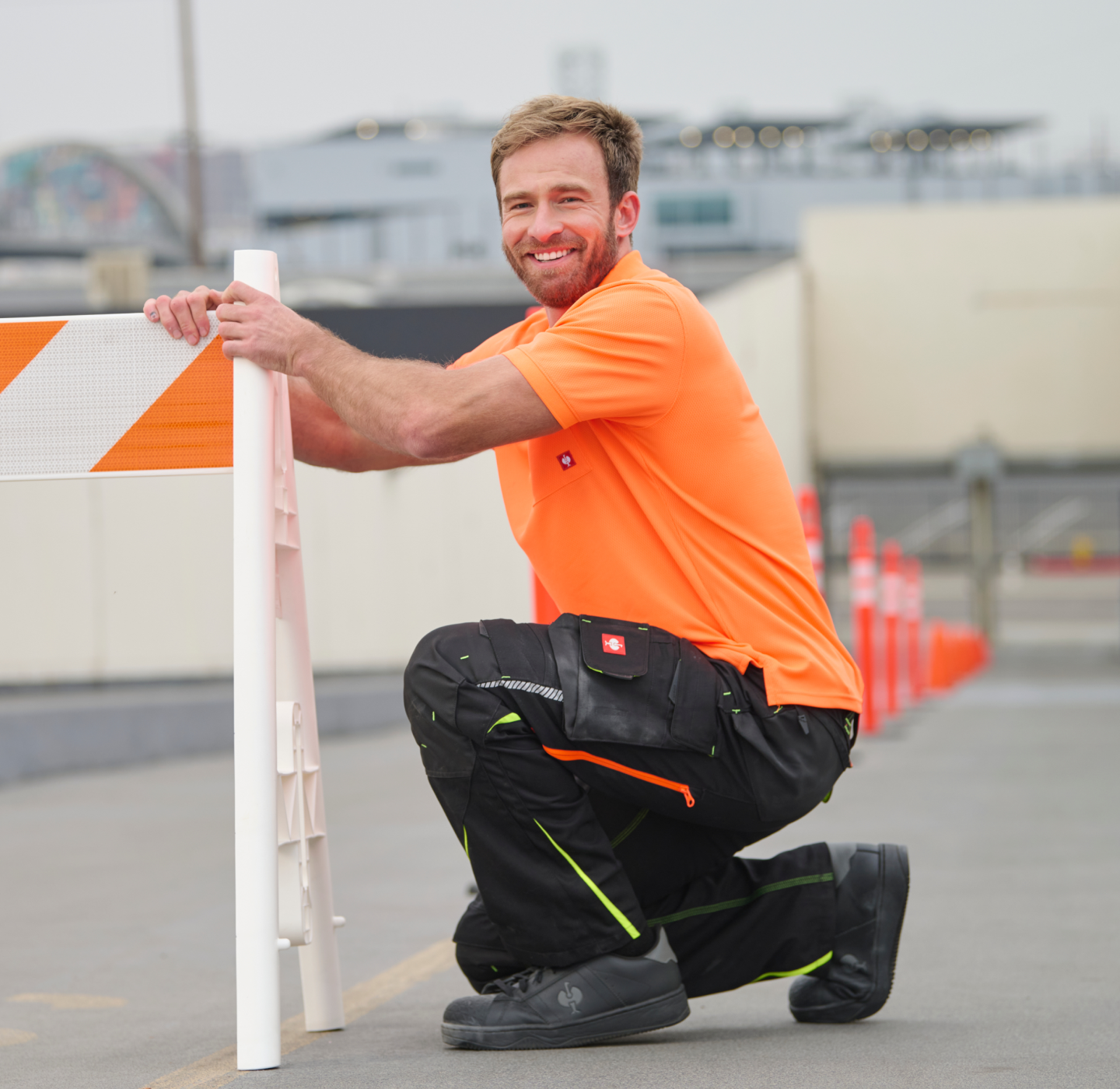 A man in an orange shirt and black work pants kneels on a street, smiling while setting up a road barrier with orange safety cones lined up in the background.