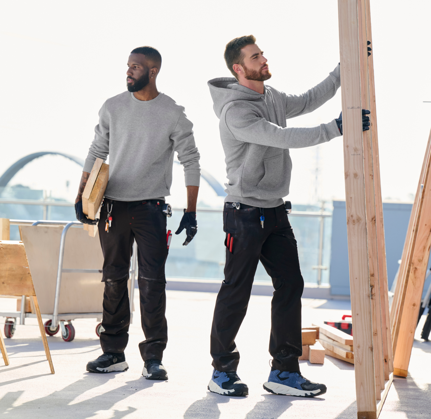 Two construction workers in hoodies and gloves work outdoors; one holds a wooden plank while the other installs a wooden frame. Tools are attached to their belts and work benches are nearby.