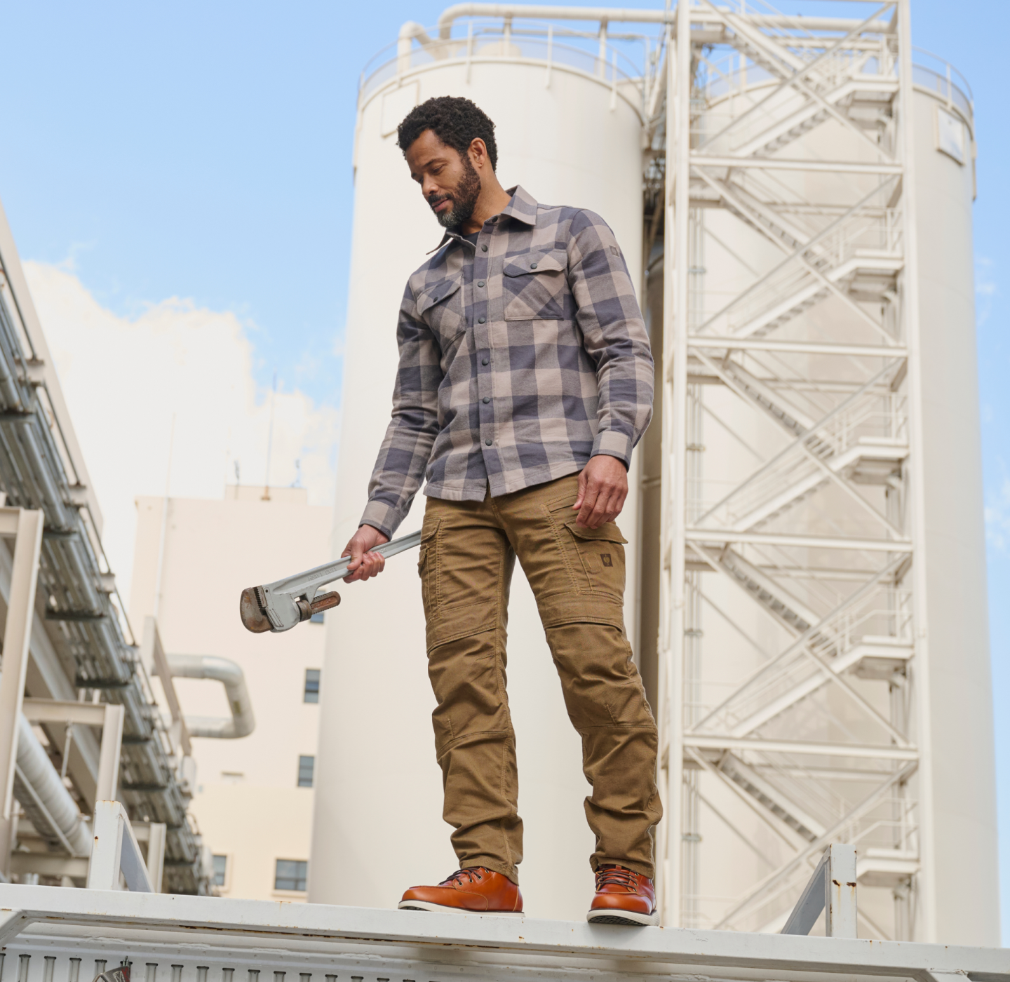 A man in work clothes holds a large pipe wrench while standing outdoors near industrial storage tanks and pipes, looking down.