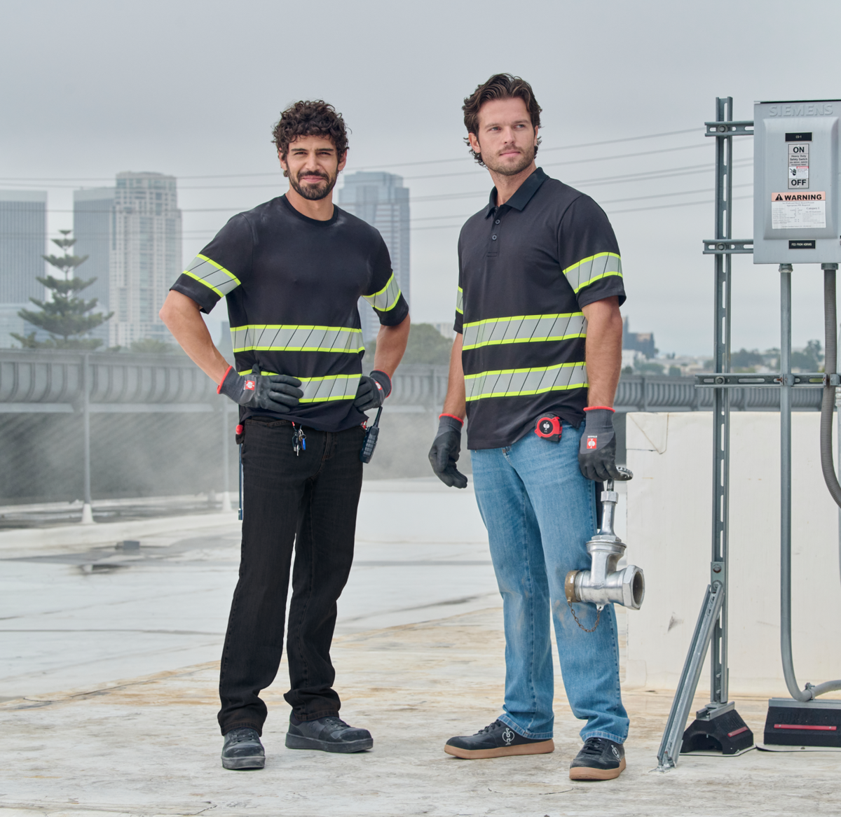 Two workers in black shirts with yellow safety stripes and gloves stand on a rooftop. One holds a pipe fitting. City buildings and electrical equipment are visible in the background under a cloudy sky.