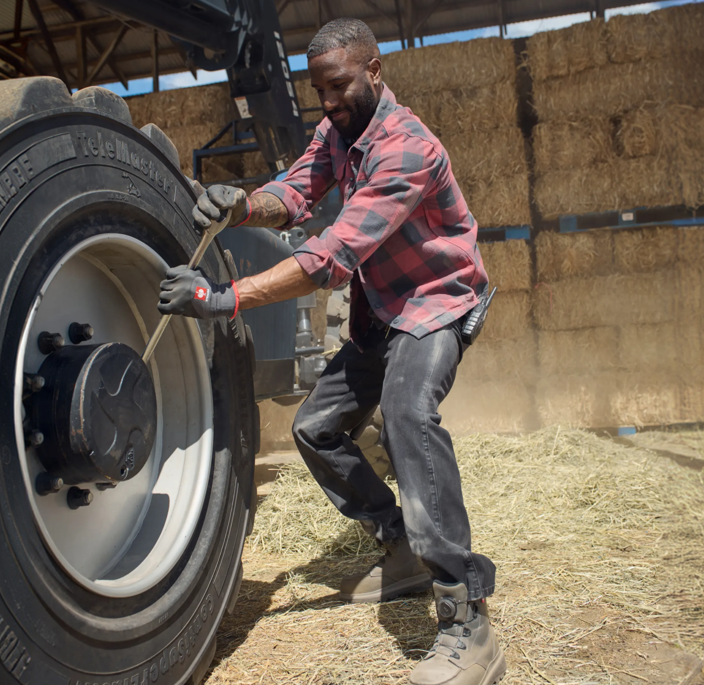 A man wearing gloves and a plaid shirt pulls a rope attached to a large tractor tire in a barn filled with hay bales.
