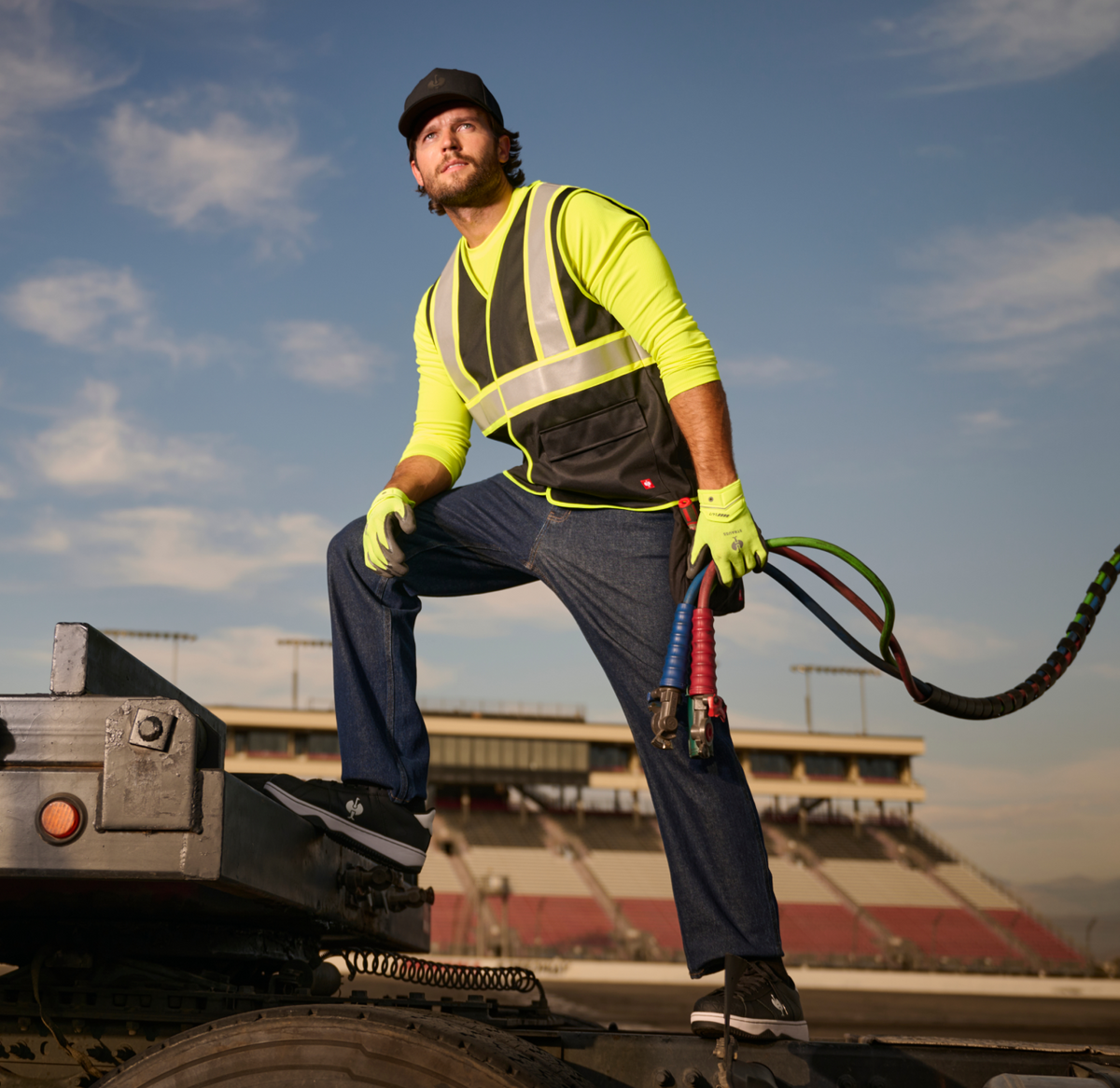 A worker in a neon shirt, safety vest, gloves, and cap stands on truck equipment, holding colorful hoses, with a stadium in the background under a partly cloudy sky.