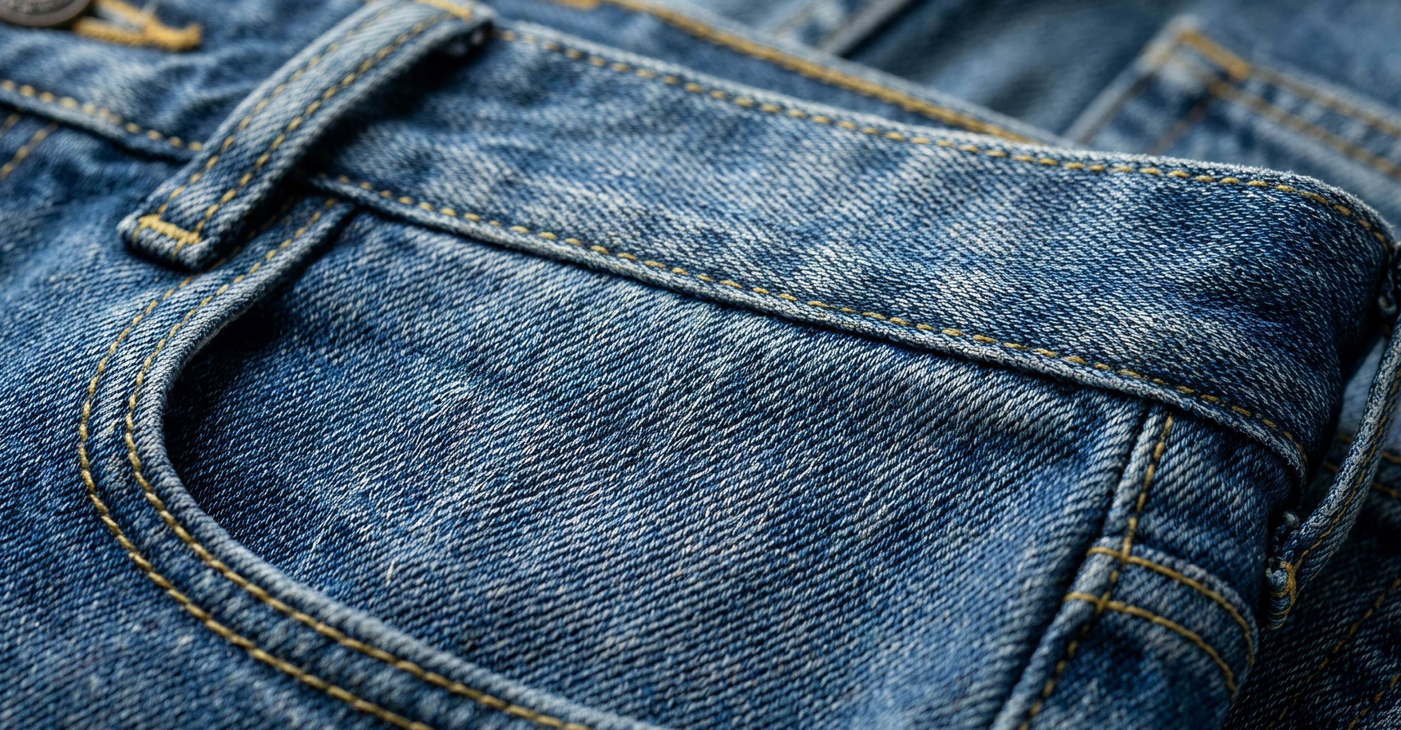 Close-up view of the front pocket and belt loop on a pair of blue denim jeans, showing the fabric texture and yellow stitching.