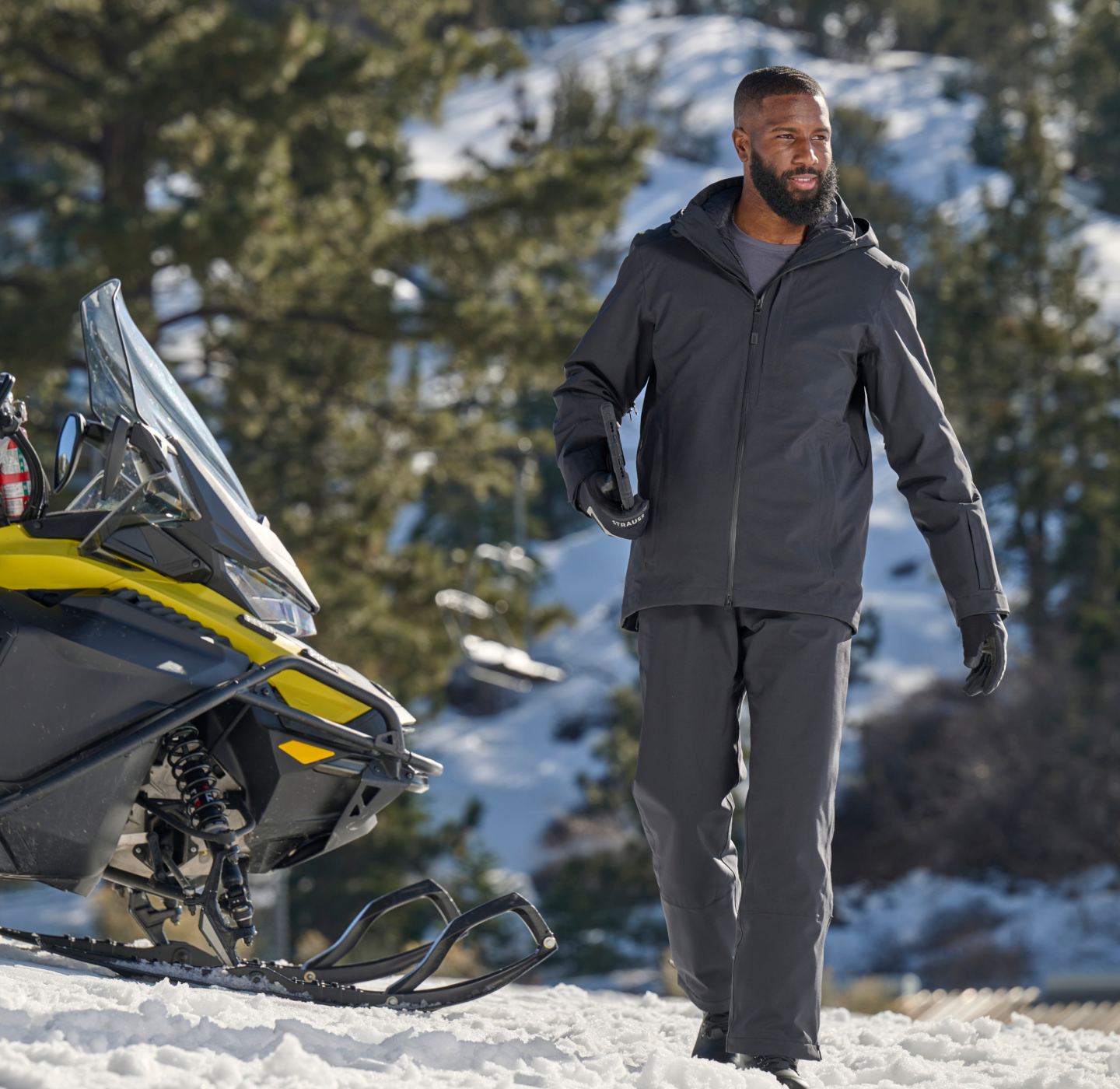 A man in a dark winter outfit stands on snow next to a yellow snowmobile, with trees and snowy hills in the background.