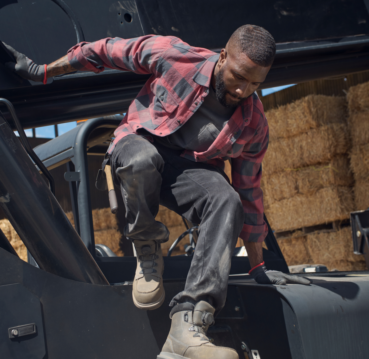 A man in a red plaid shirt climbs down from farm equipment, with hay bales stacked in the background.