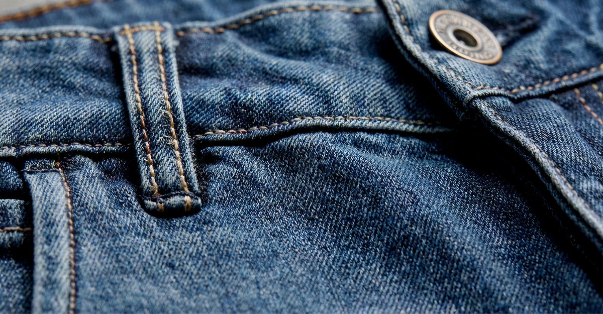 Close-up of blue denim jeans showing a belt loop, seam, and a metal button on the waistband. The fabric texture and stitching details are clearly visible.