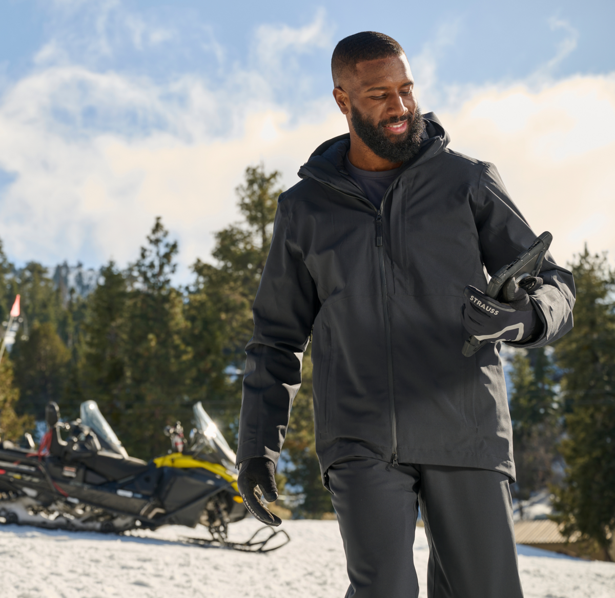 A man in winter clothing stands on snow, smiling and putting on gloves. Snowmobiles and evergreen trees are visible in the background under a partly cloudy sky.