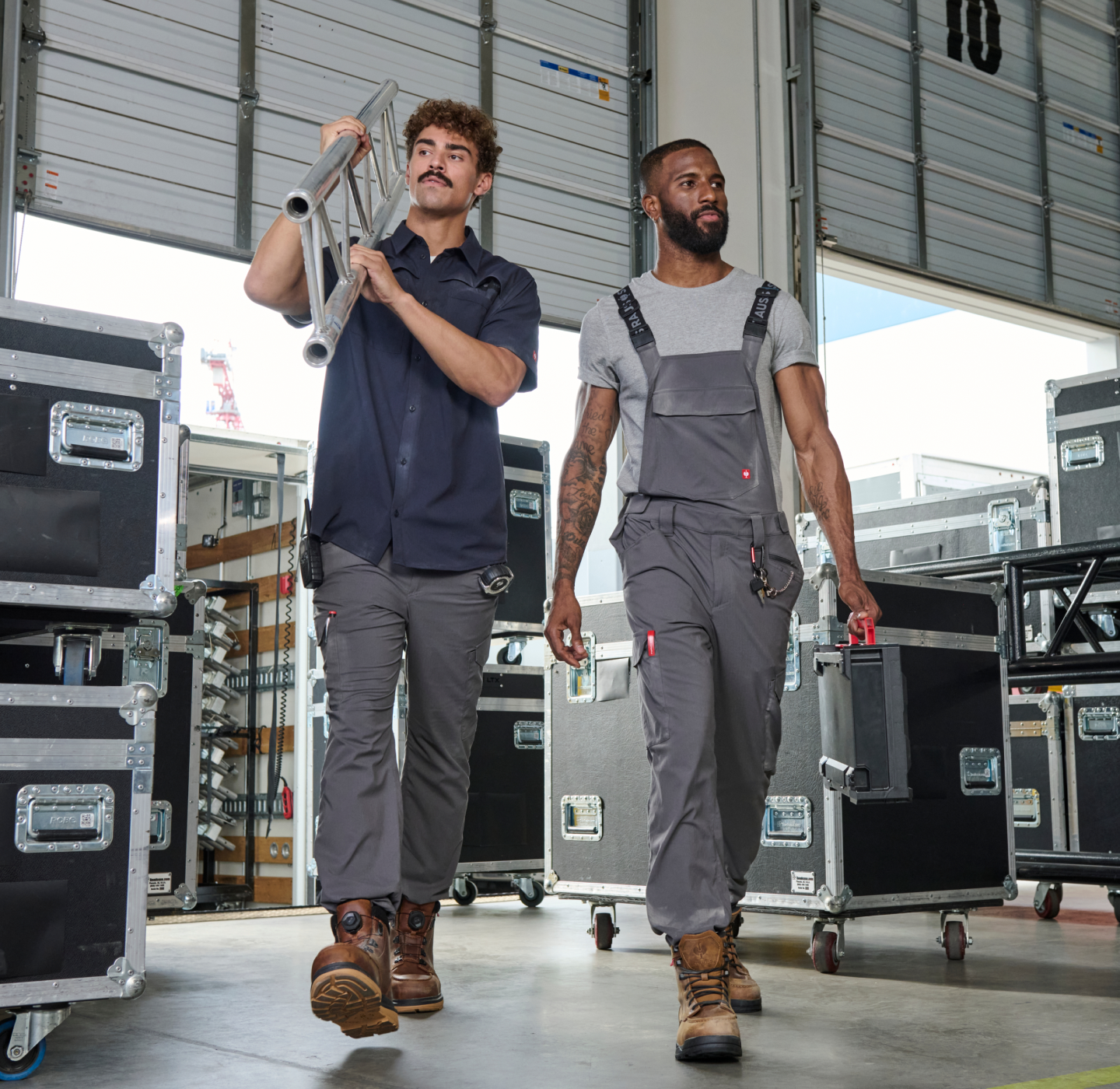 Two men in work attire walk through a warehouse filled with equipment cases. One carries a folded ladder, the other holds a toolbox. Large open garage doors and bright natural light are visible in the background.