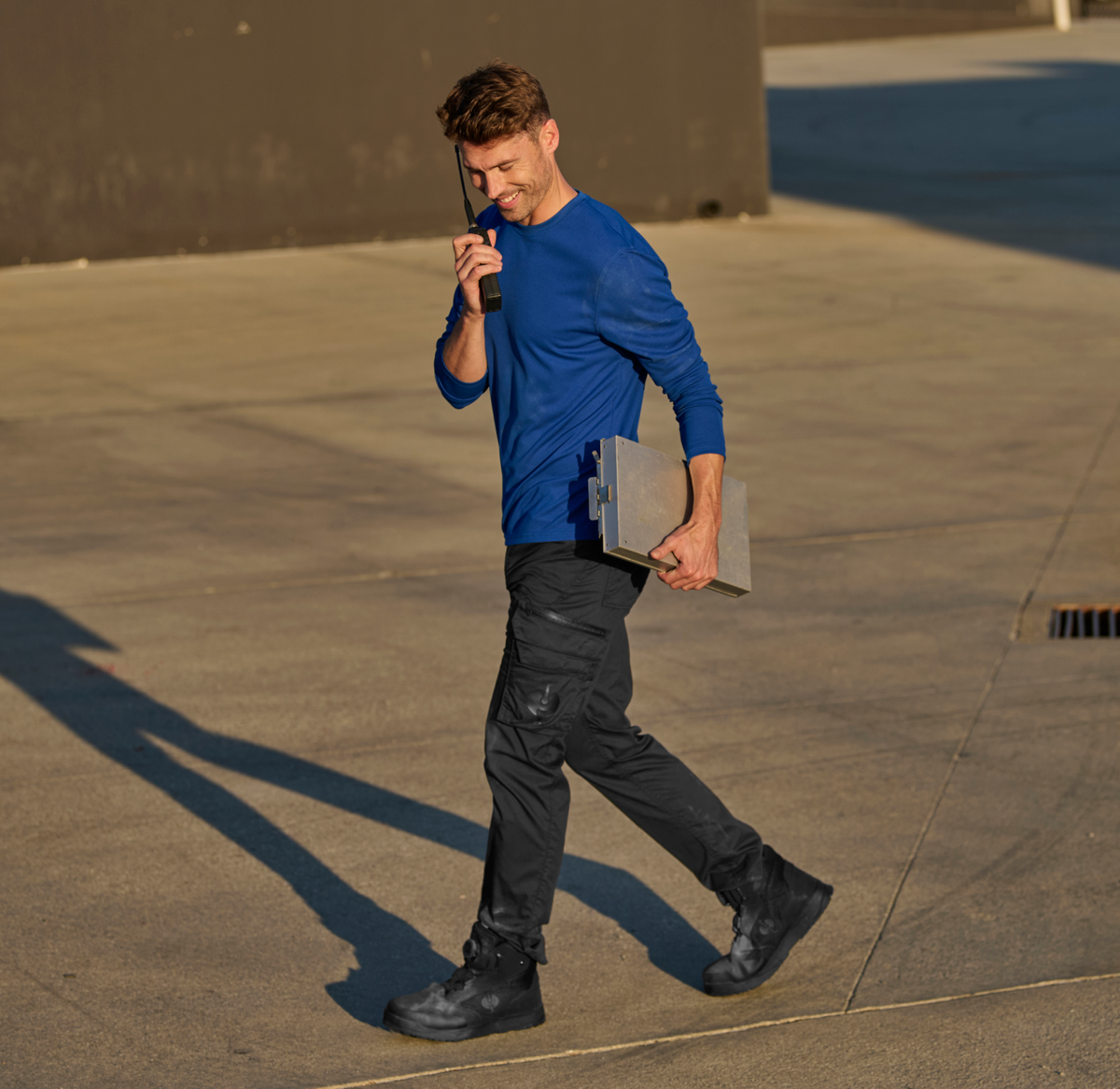 A man in a blue shirt and black pants walks outside on concrete, holding a clipboard and talking on a walkie-talkie, smiling. Shadows and sunlight are visible on the ground.