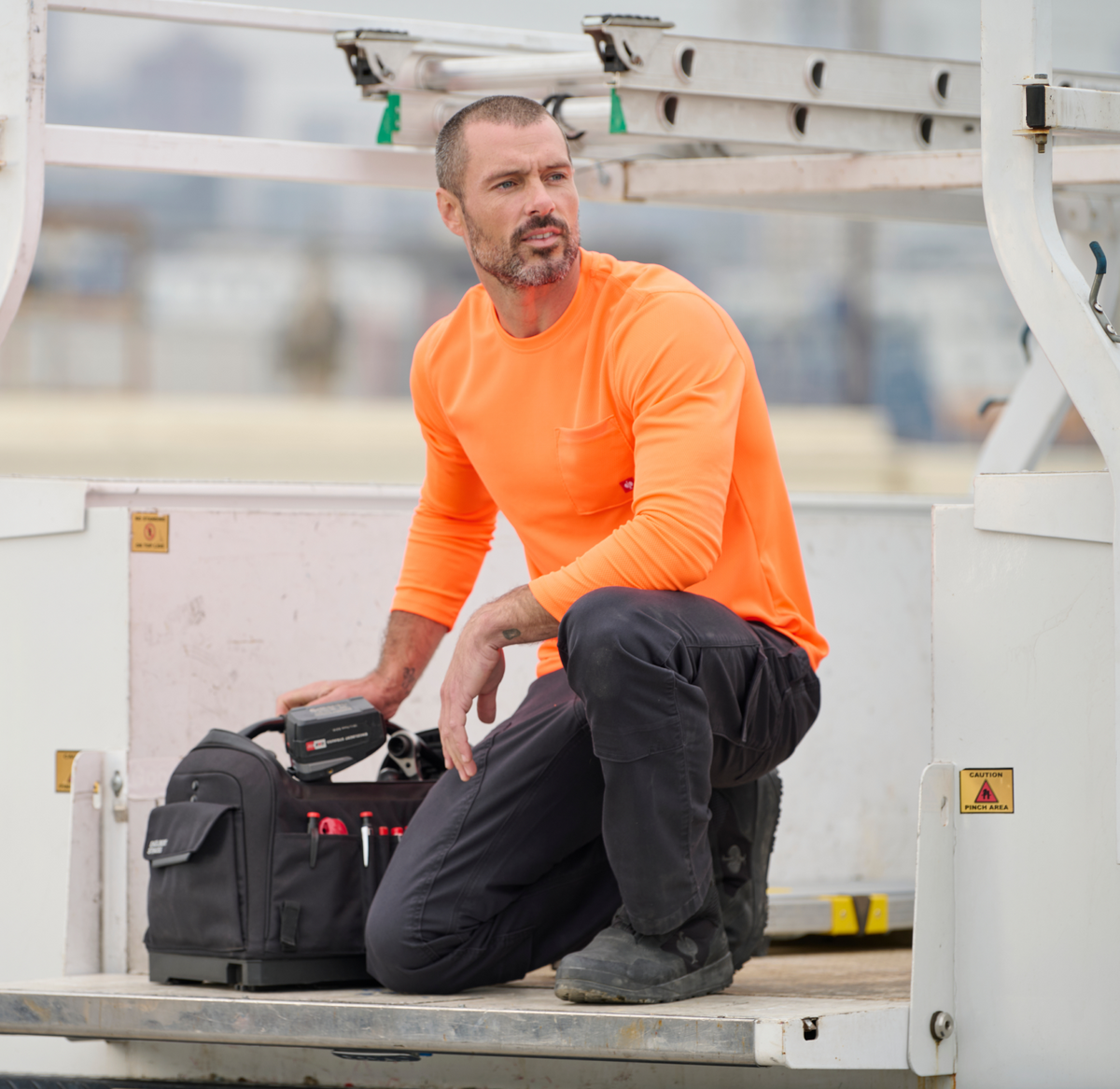 A man in a bright orange long-sleeve shirt kneels on a platform, holding a tool near a black tool bag, with industrial equipment in the background.